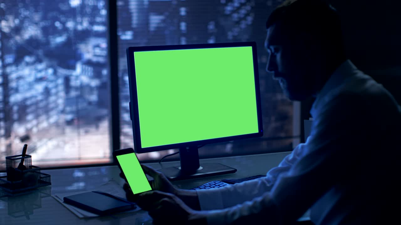a última hora de la noche, el empresario trabaja en una computadora personal y un teléfono inteligente con pantalla verde en su oficina privada con vista a la ventana de la gran ciudad.
