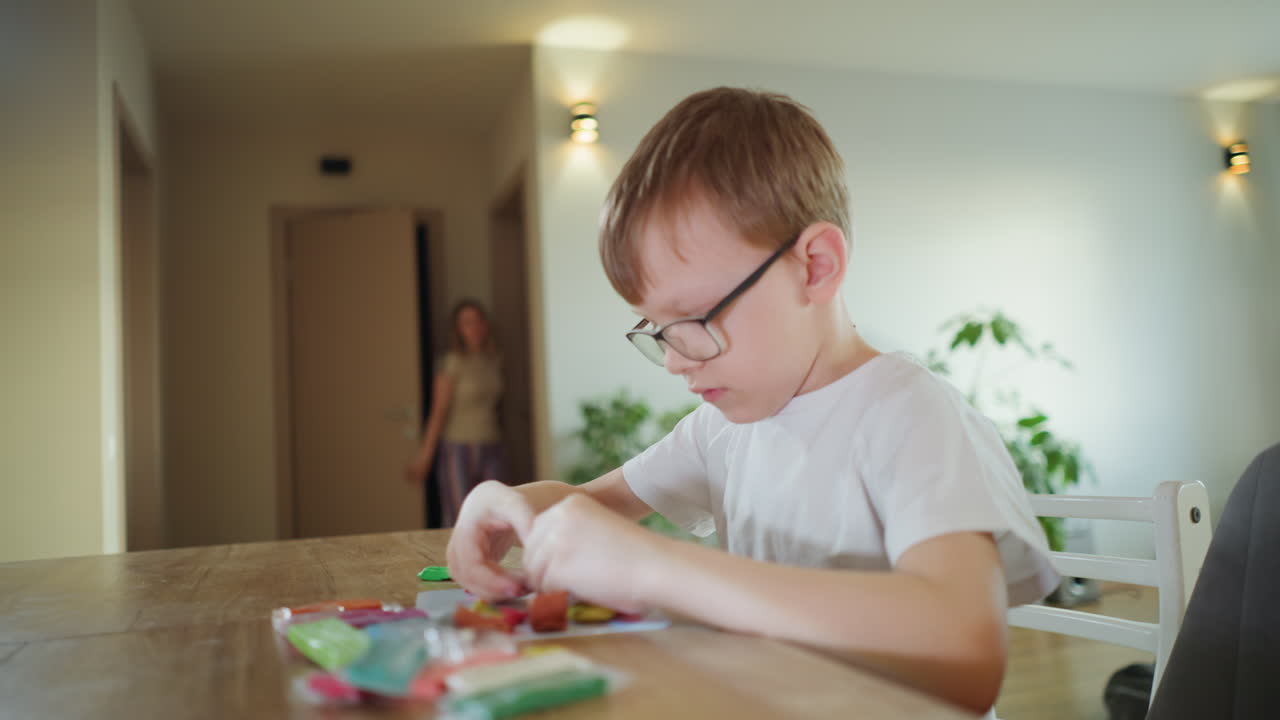 Boy wearing glasses sits at wooden table shaping colorful clay pieces with concentration, engaging in creative hands-on activity while crafting small figures, fine motor skills, focus at home