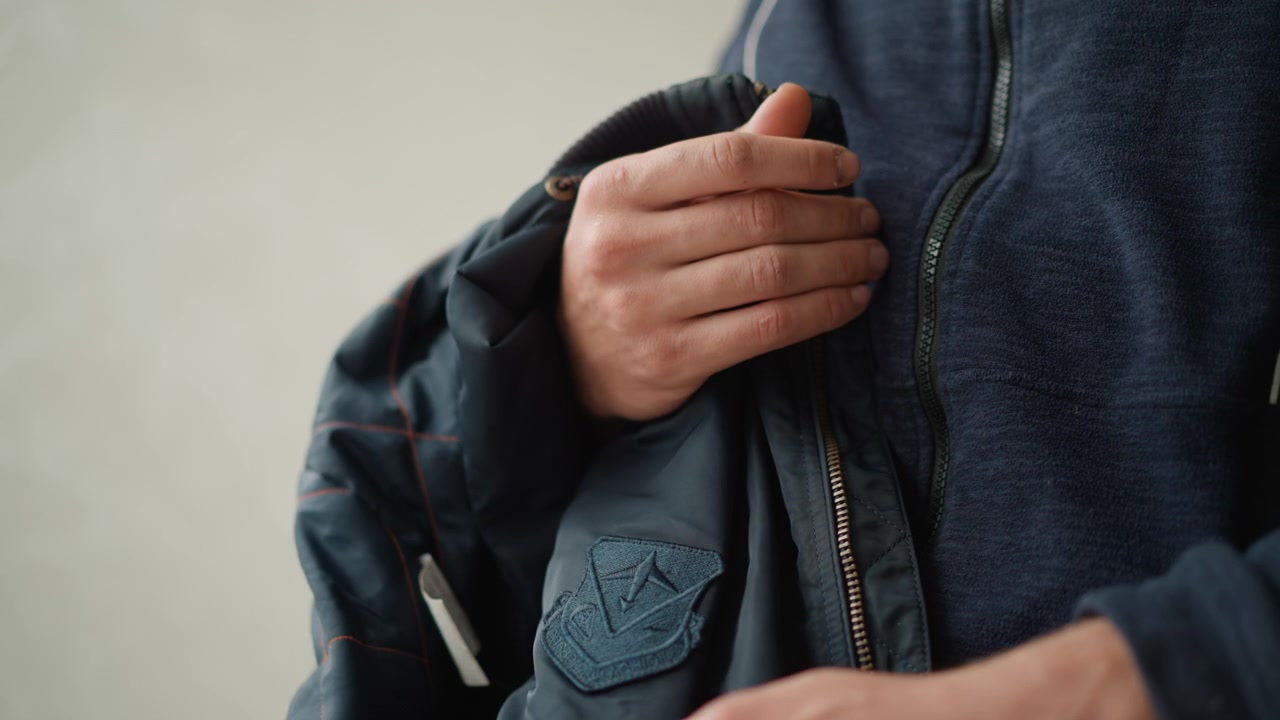 Close up of man holding and adjusting his dark blue jacket with visible stitching details. focus remains on hands fabric texture zipper pull and embroidered patch throughout sequence. Soft