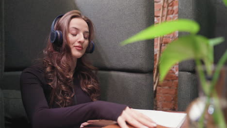 light skin lady with blond hair wearing headset and smiling gently while seated in cozy booth. she appears relaxed and immersed in sound, enjoying peaceful moment with notebook on table nearby