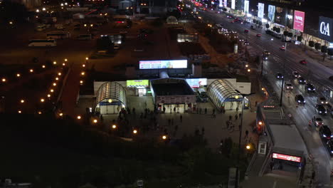 Slide-and-pan-footage-of-illuminated-Metro-entrance-near-wide-multilane-street-at-night.-People-walking-from-subway.-Warsaw,-Poland