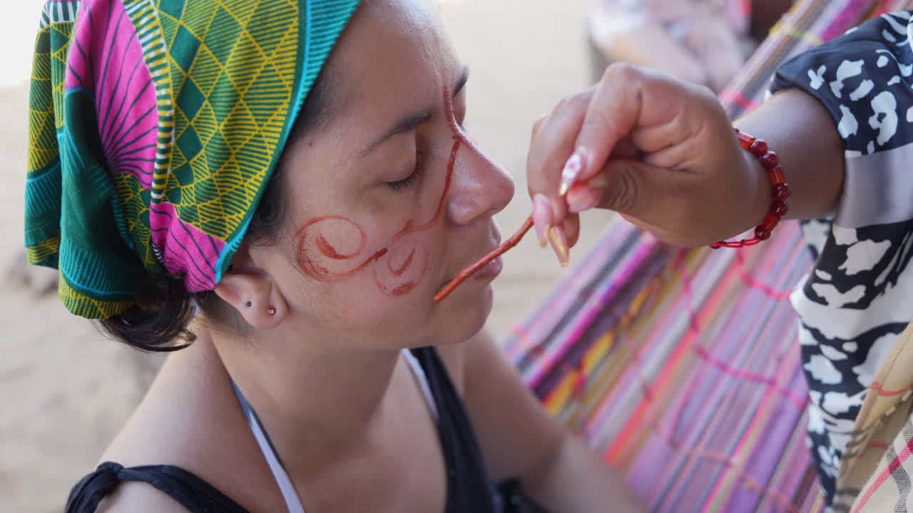 Nahaufnahme von Wayuu-Frauen in La Guajira, Kolumbien, die traditionelle Gesichtsbemalung mit natürlichen Pigmenten auftragen. Lebendige Textilien, ethnische Muster und natürliches Tageslicht unterstreichen die kulturellen Details.