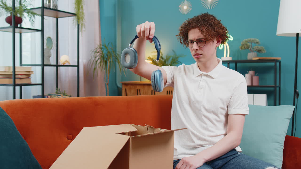 불행한 남자가 상자를 열고 손상된 무선 헤드폰을 집에 가져오는 것에 불만을 느니다.
