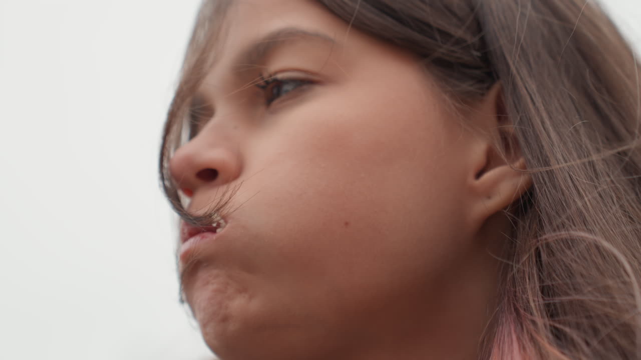 Una studentessa latina adolescente mangia un panino all'aperto, con la bocca sporca di briciole, i capelli scompigliati dal vento, cielo nuvoloso, ritratto ravvicinato che mostra un'espressione pensierosa e un'azione spontanea del masticare, cappotto casual e plastica