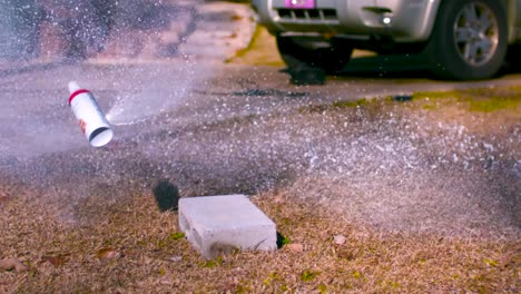 a small bullet being shot through a whipped cream can under pressure and whipped cream starts foaming and blowing out of two bullet holes during a summer day in a garden, with a car in the background.
