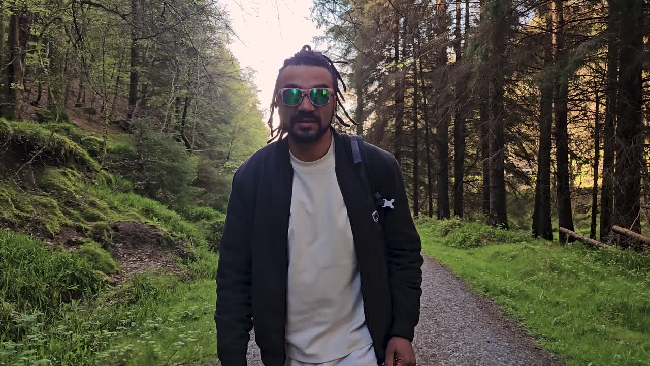 Sort mand kigger lige frem, mens han kommer mod kameraet ad en jordsti i en grøn efterårsskov. Kameraet bevæger sig baglæns med dolly.