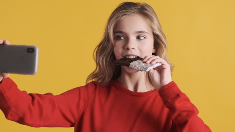 teenage caucasian girl eating chocolate bar and taking selfies with smartphone.