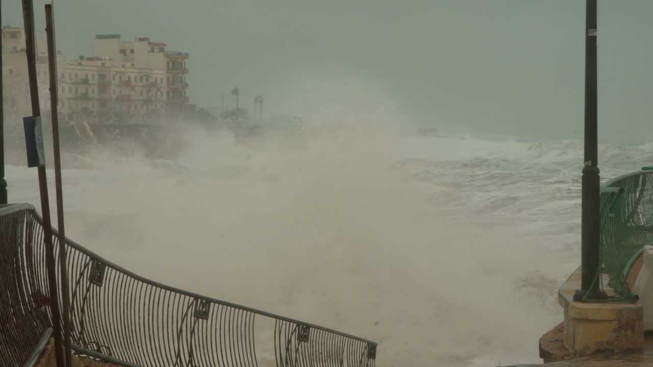 Slow-motion shot of a stormy scene in Marsaskala, Malta, during Storm Harry, captures rough seas crashing against a damaged promenade under overcast skies, showcasing extreme winter weather