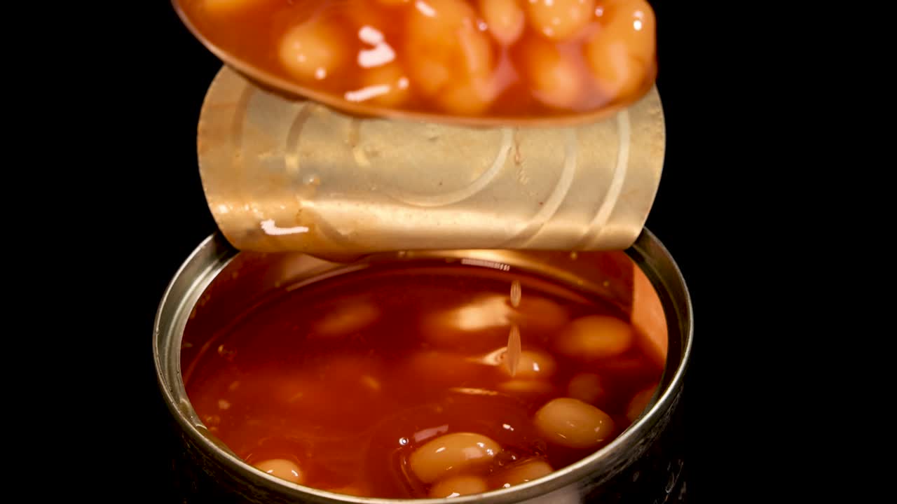 A metal spoon lifts baked beans in tomato sauce from an opened can against a black background. Overhead close-up, even lighting, minimal shadows