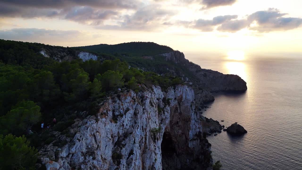 Escursionisti riposano su una scogliera rocciosa tra gli alberi con vista sul paesaggio verde durante il tramonto a Ibiza