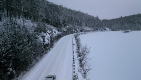 an aerial drone view of a car driving along a snowy winter country road between a frozen lake and forest scene