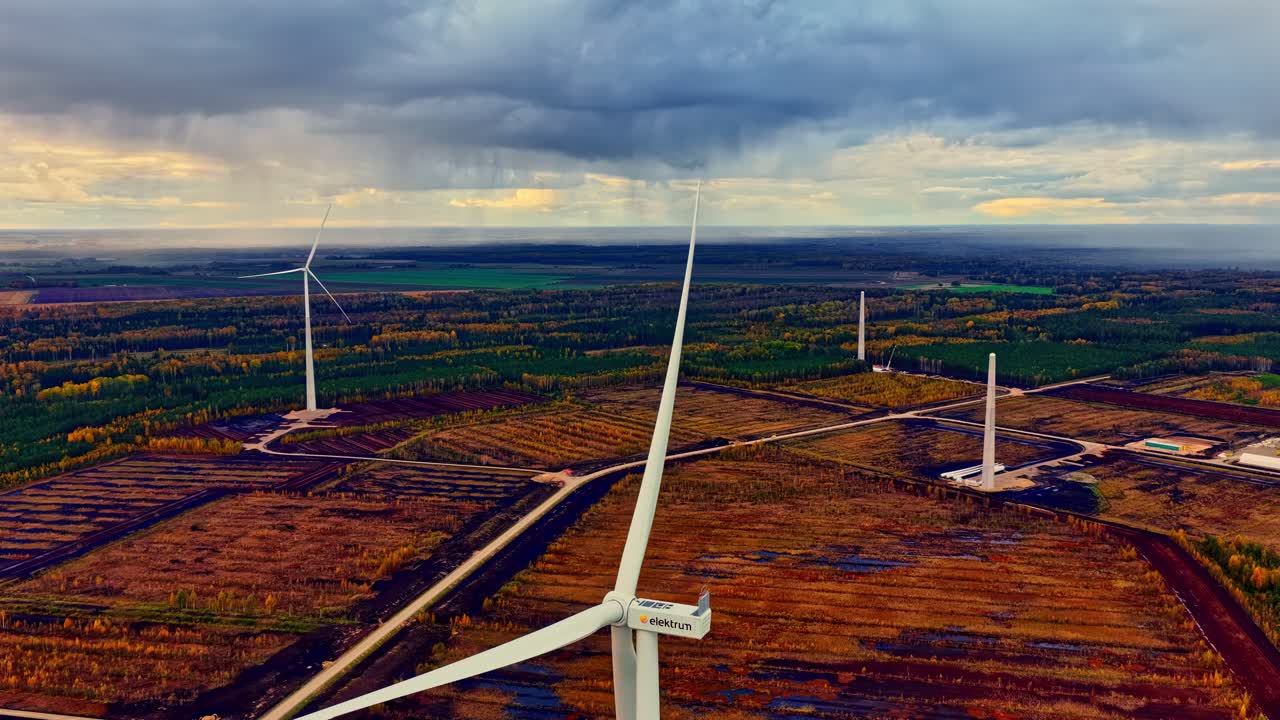 Aerial tracking shot above rotating wind turbine blades across green fields and distant forest in soft light