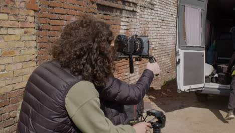 Close-Up-View-Of-Redheaded-Production-Worker-In-Wool-Cap-And-Sunglasses-Setting-Up-A-Camera-In-The-Street-While-Another-Coworker-Helping-Him