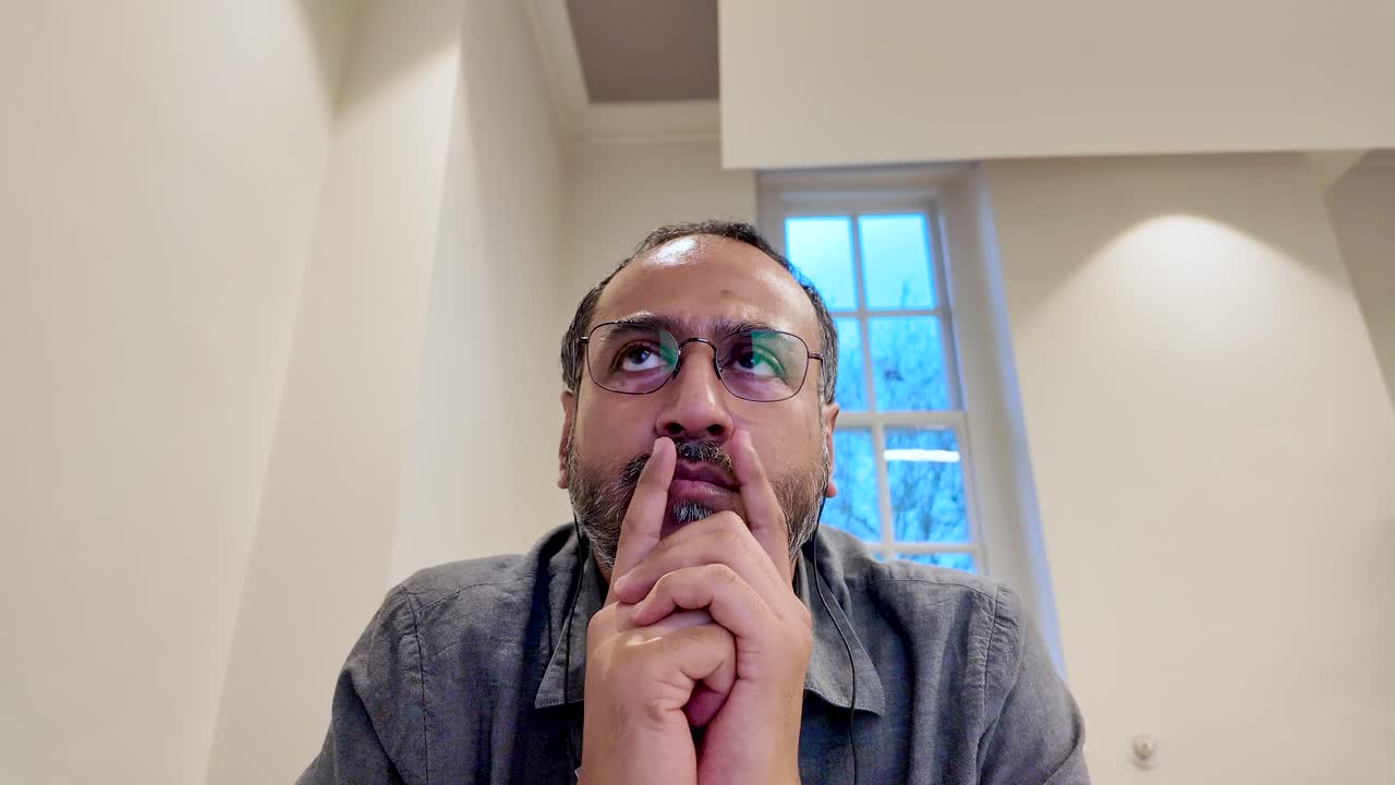 Pensive South Asian Man With Hands On Lips Wearing Glasses Deep In Thought Contemplating Ideas Problem Solving Brainstorming Solutions Indoors