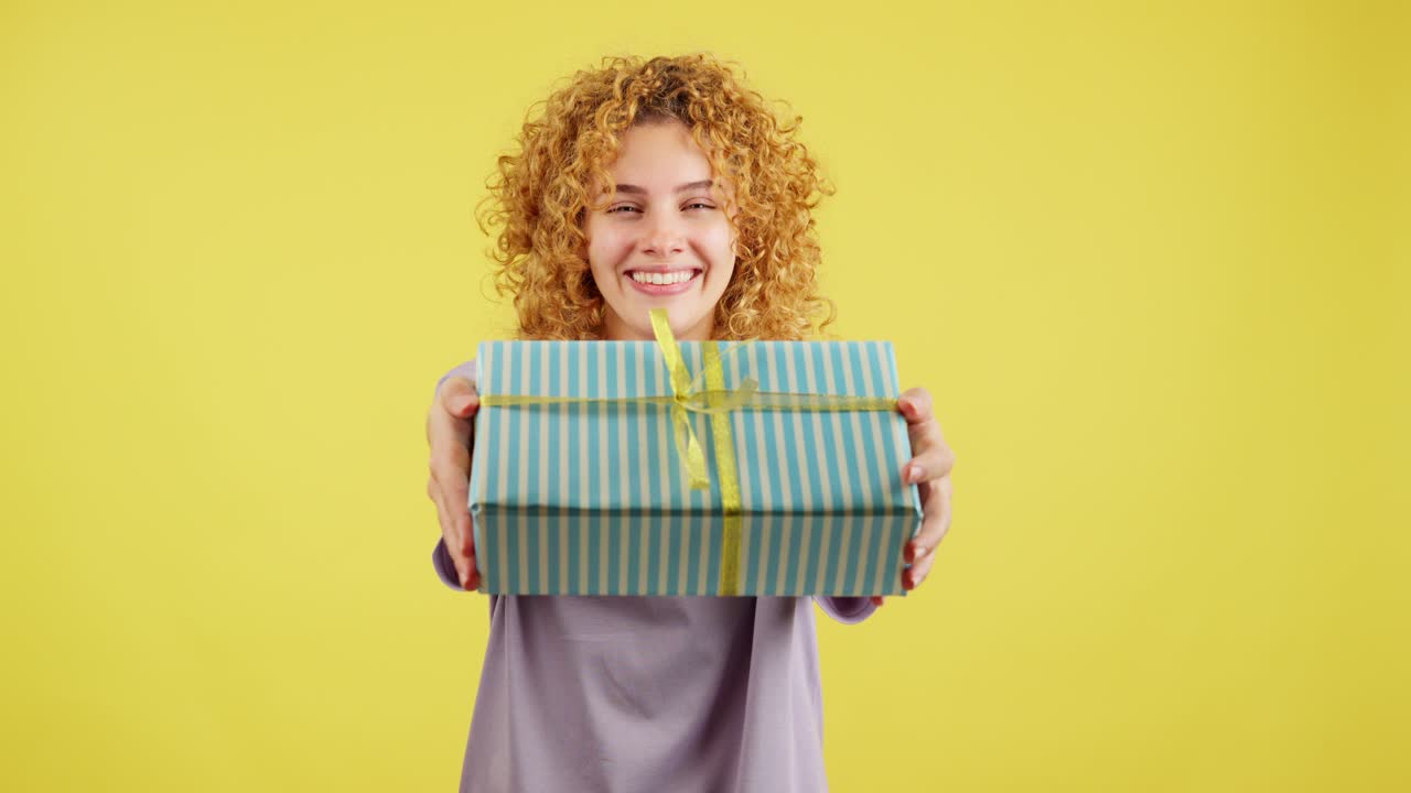 A cheerful woman with curly hair holding out a striped gift box on a yellow background