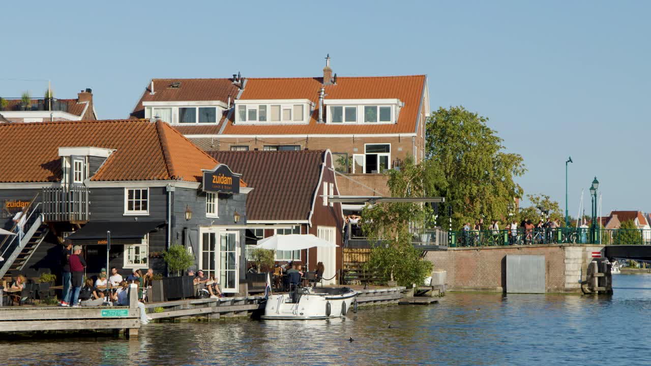 People gather at waterfront bar and restaurant beside canal, bright daylight, steady wide shot