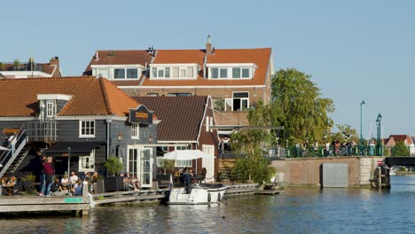 people gather at waterfront bar and restaurant beside canal, bright daylight, steady wide shot