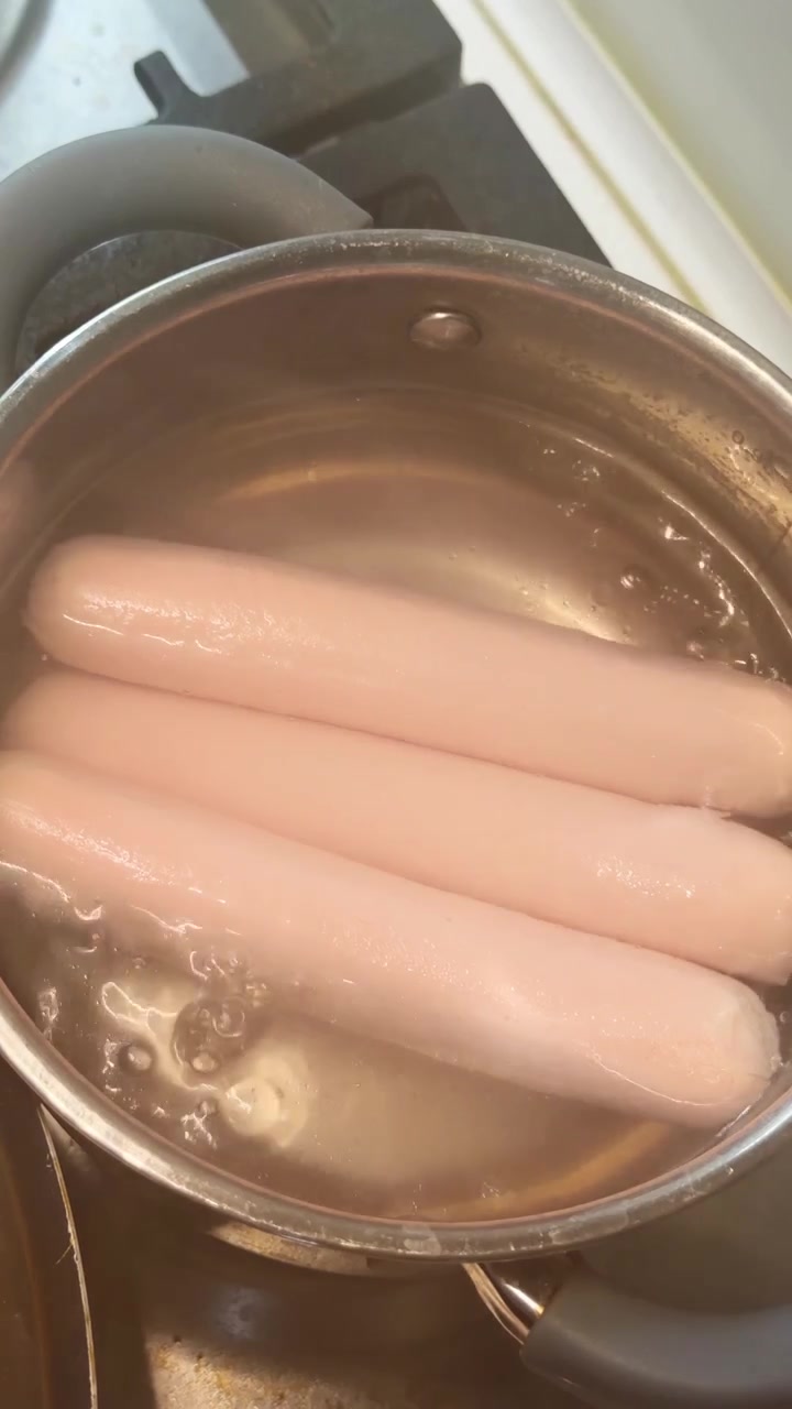 Salsicce che bollono in acqua all'interno di una pentola in acciaio inossidabile. Tre würstel rosa immersi in acqua che sobbolle all'interno di una padella metallica sotto una luce da interni