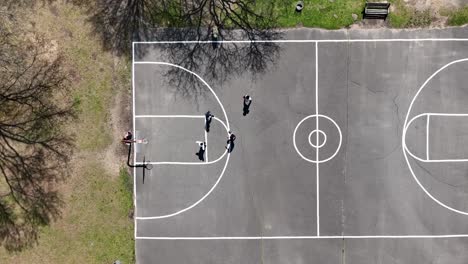 An-aerial-view-over-four-people-playing-half-court-basketball-surrounded-by-dry-trees-on-a-sunny-day