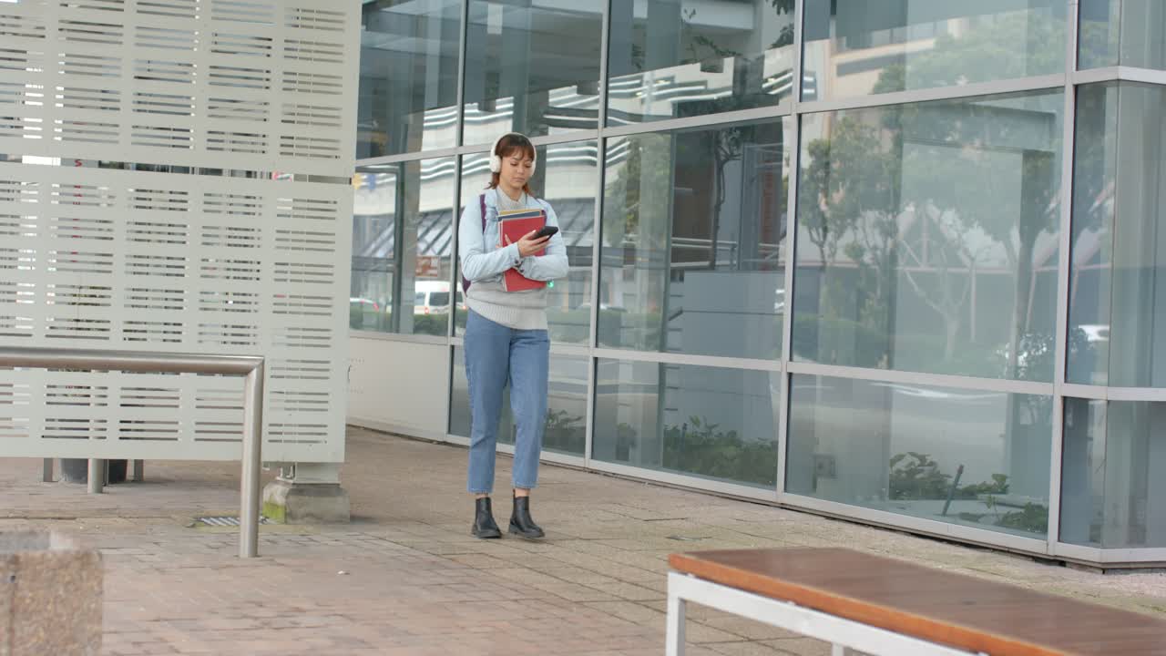 Una donna entra nell'inquadratura, cammina lungo un edificio di vetro, tiene una cartella rossa e tocca lo smartphone, con spazio per il testo.
