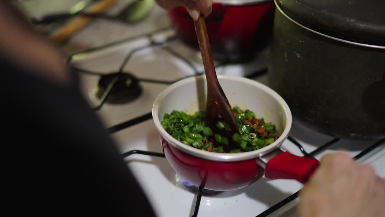 Person stirring chopped fried sauce vegetables for Locro Argentino in pot over gas stove in rustic kitchen.