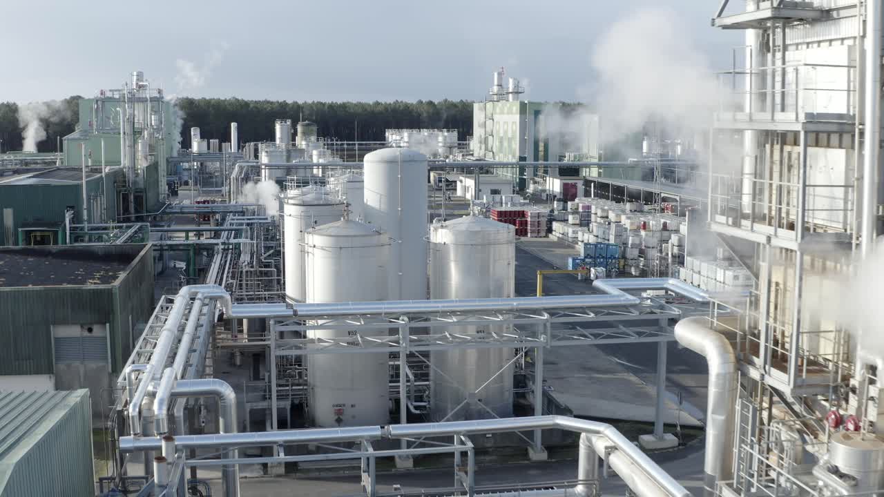 Aerial drone navigates through sections of large factory, highlighting storage tanks, extensive pipe networks, and steam emissions, Castets, France