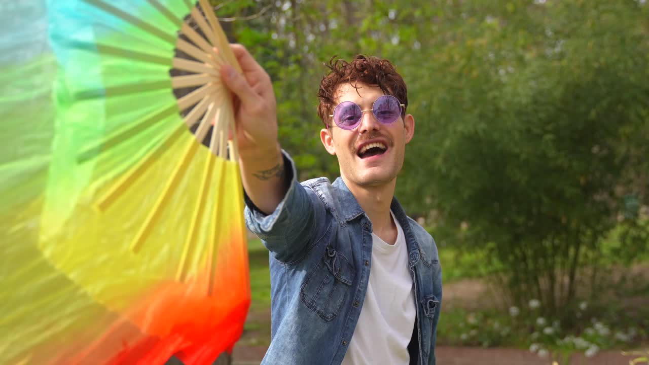 Um homem posa com um leque com as cores do arco-íris em um parque