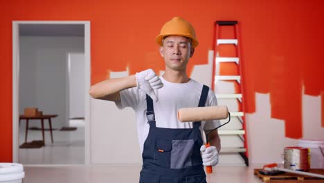 pintor asiático con casco de seguridad mostrando los pulgares hacia abajo gesto a la cámara mientras está de pie con la pared de pintura en la habitación
