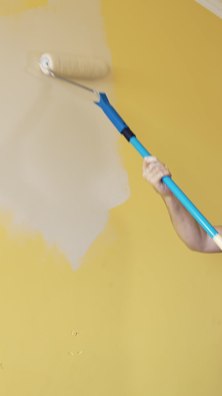 Room improvement with a new color. Professional worker in blue overalls painting wall with a paint roller on a long handle on. Craftsman working. Vertical video