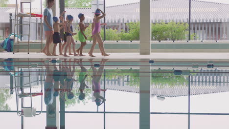 male coach walks children in swimming class along edge of indoor pool