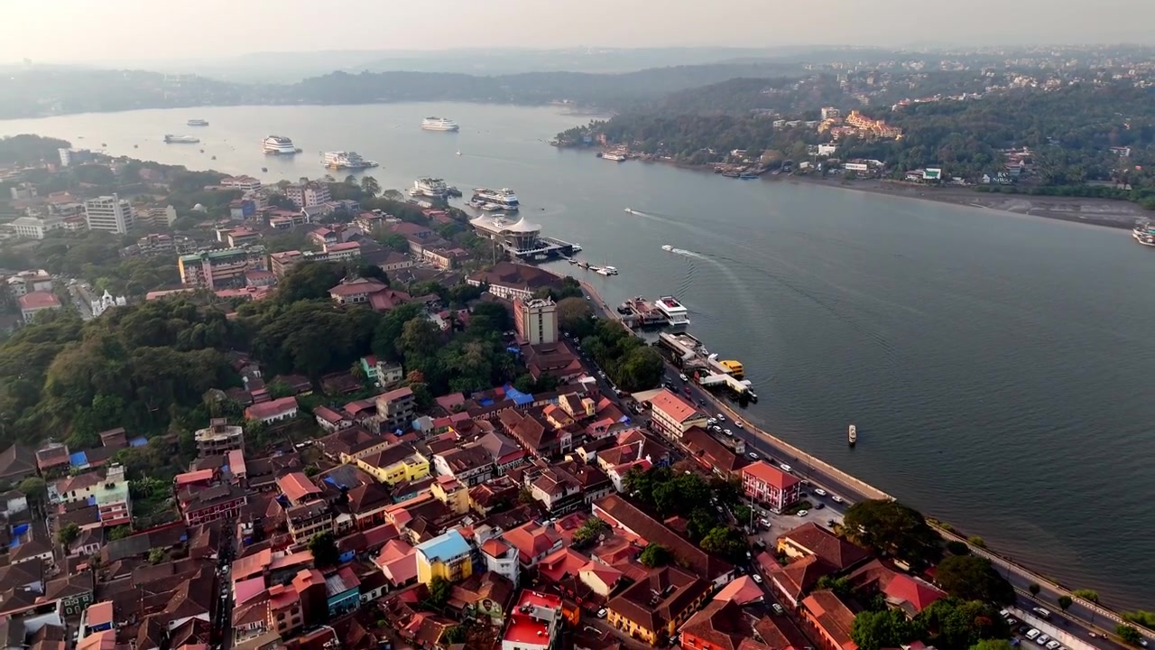 Veduta aerea cinematografica del fiume Mandovi che mostra iconiche navi casinò ancorate lungo la riva, con barche in movimento che creano increspature sulle acque calme ed edifici cittadini che si affacciano sulla sponda.