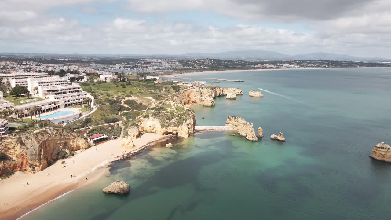 Aerial drone shot shows sandy beach, rugged cliffs, turquoise water, and Lagos buildings in Algarve
