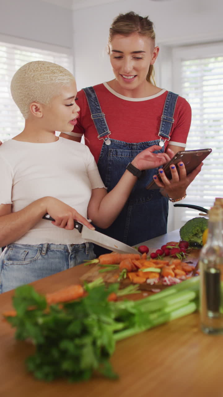 video vertical de una feliz pareja de mujeres diversas abrazando y usando la tableta en la cocina