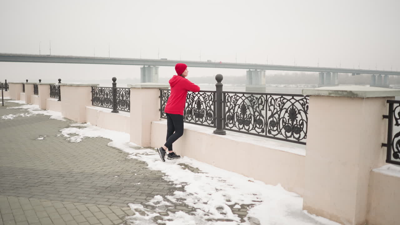 Athlete in red hoodie and black leggings walks to decorative fence, resting hand on rail with gloves, gazing at distant bridge with moving cars in misty winter atmosphere, snow covering ground