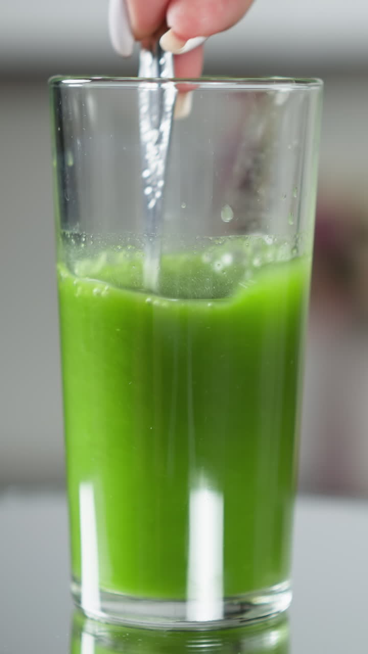 una mano delgada sostiene una cuchara en un vaso lleno de jugo de verduras rico y verde