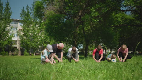 grandparents and their grandsons are all kneeling on a grassy field, tying their shoelaces, the youngest boy is resting his head on a soccer ball, with his brother in red looking at him