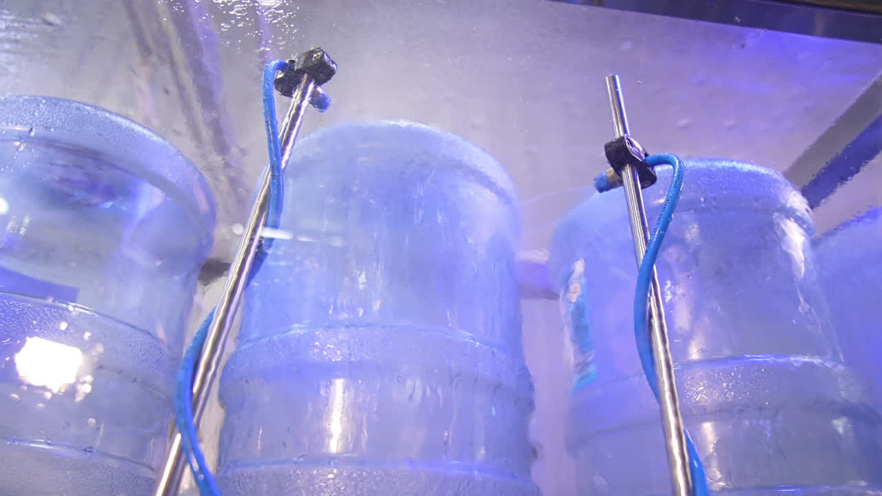 Blue big bottles are put upside-down and being washed. Flasks are sprayed with water for cleaning. Low angle view.