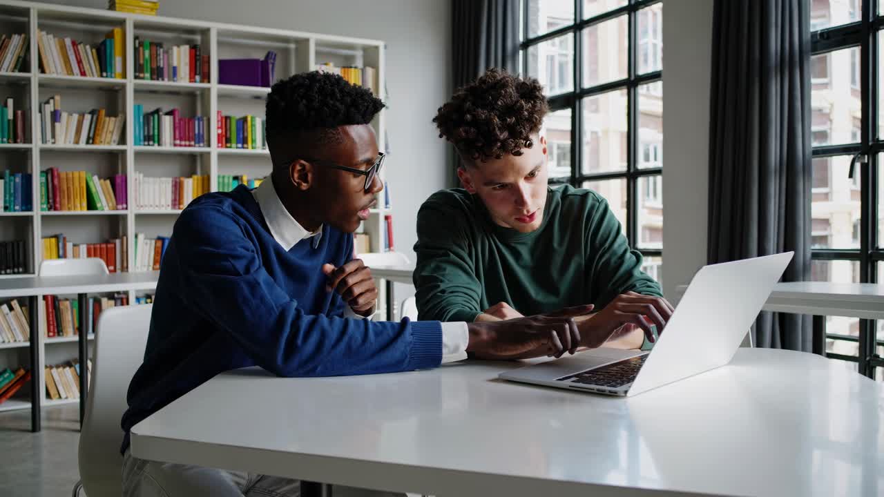 Two men discuss at a table with a laptop, framed by bookshelves. The video captures a side angle