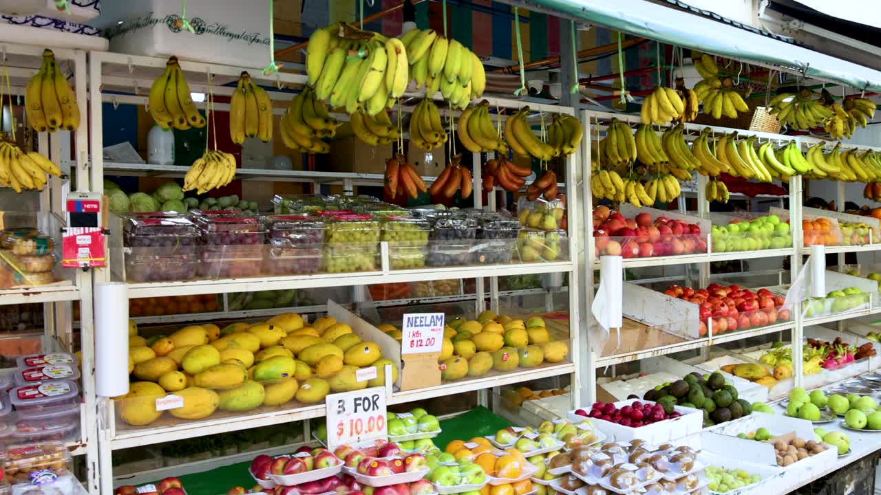 Vendor arranges bananas, mangoes, and fresh fruit at colorful outdoor market stall in daylight