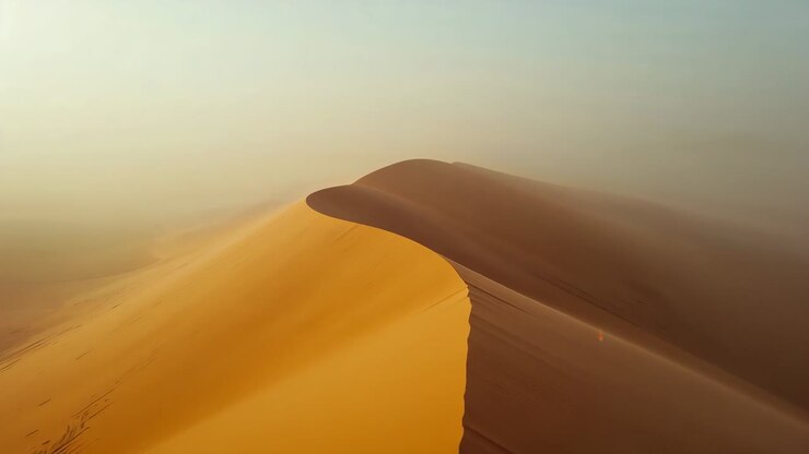 Gliding aerial camera following dune crest in desert, wind lifting sand revealing ripples