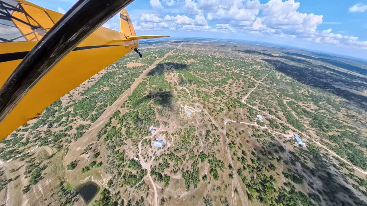 POV aerial footage from a yellow light aircraft flying over a rural landscape. The clip shows the aircraft wing, its shadow on the ground, winding dirt roads, scattered trees, and open fields