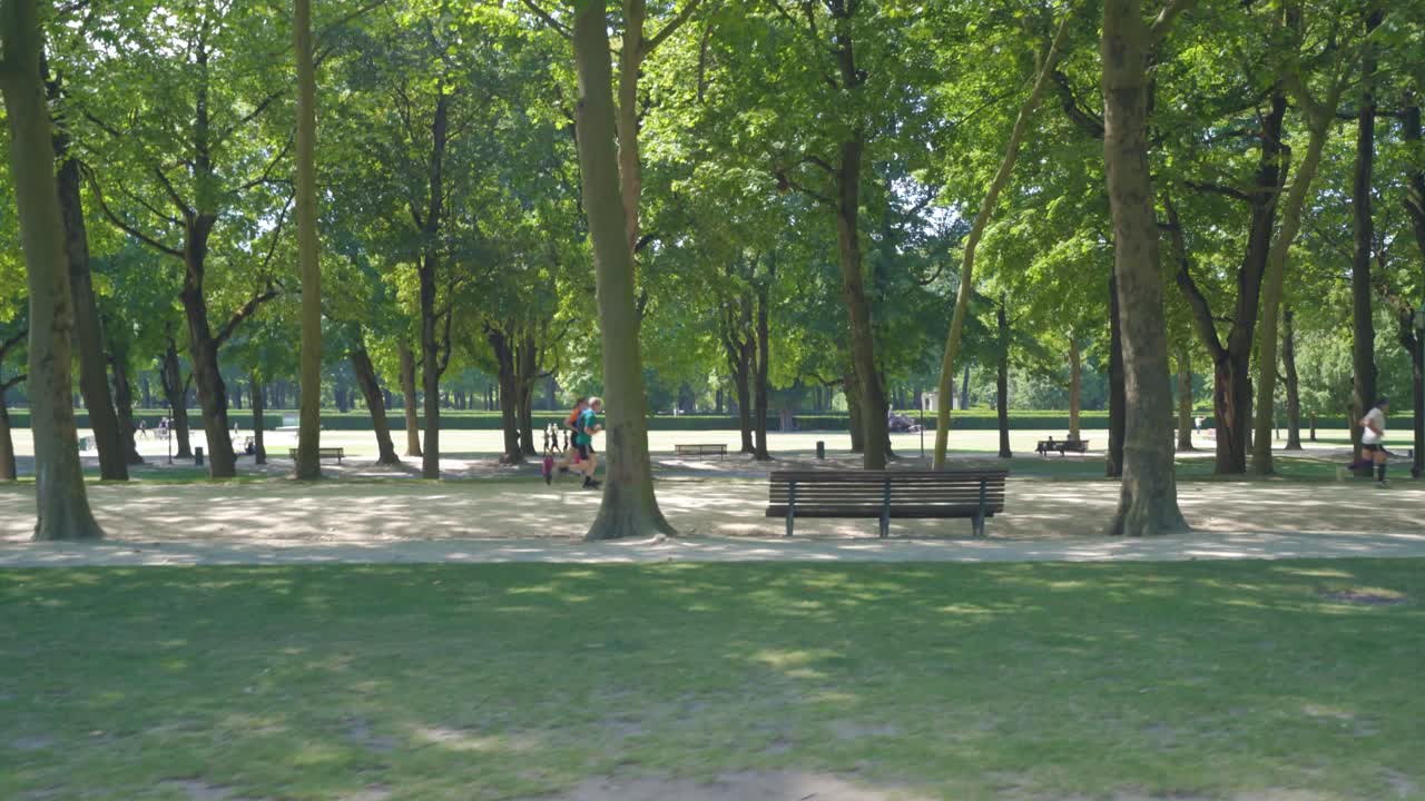 Sideways Dolly shot of two runners on park way with green trees and foliage on side of park street. Warm sunny summer day in the park of Cinquantennaire park in Brussels, Belgium, people in distance.