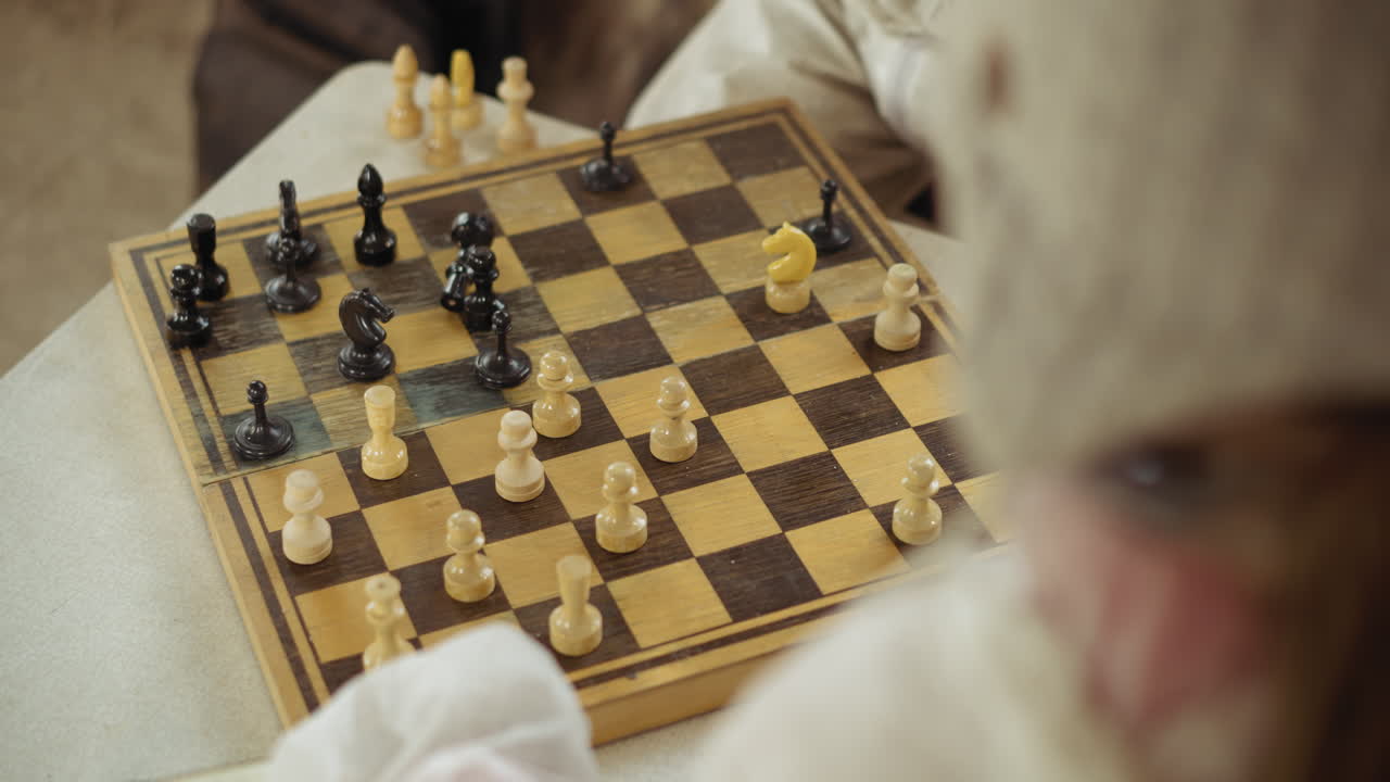 Rear view of focused person dressed in light clothing playing chess using white pieces on rustic wooden board, game is in progress as individual observes strategy