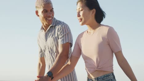 Feliz-Pareja-Birracial-Caminando-Y-Tomándose-De-La-Mano-En-La-Playa,-En-Cámara-Lenta