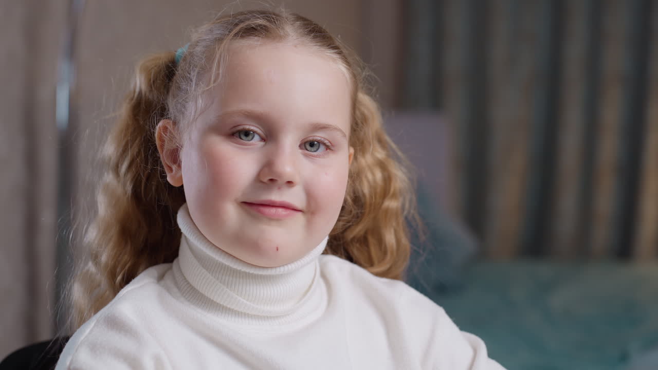 Ragazza con capelli biondi ricci raccolti in codini che indossa un dolcevita bianco e sorride dolcemente alla fotocamera in interni, illuminazione calda con sfondo sfocato che crea una calma atmosfera di innocenza infantile