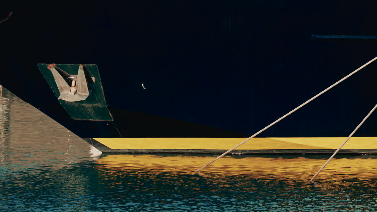 Close up of a yacht's bow showing an anchor and yellow hull reflection shimmering on calm marina water