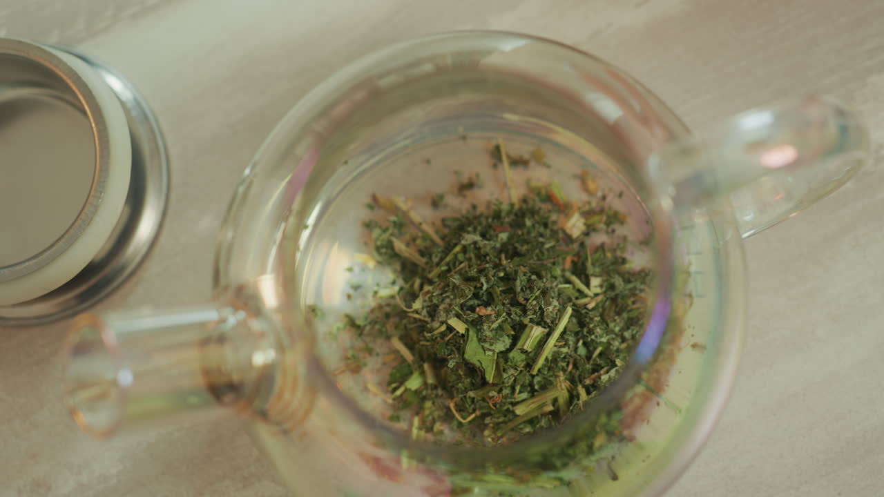 teiera di vetro con erbe vista dall'alto, mani che posizionano il coperchio e versano acqua calda sulle foglie sfuse per un'infusione delicata, vapore che sale, calda luce naturale, accogliente ambiente cucina, rituale rilassante per il benessere