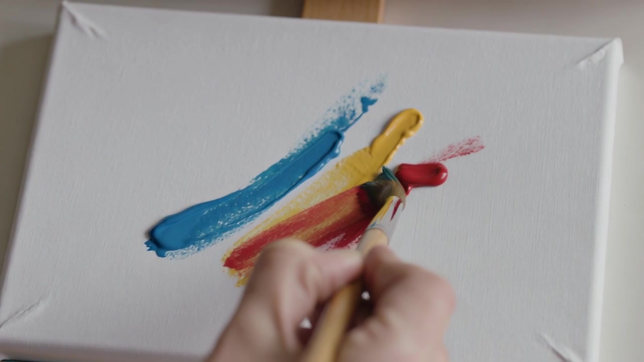 Abbassando un pennello piatto, una mano stende la pittura sulla tela su un piccolo cavalletto, mescolando giallo e rosso.
