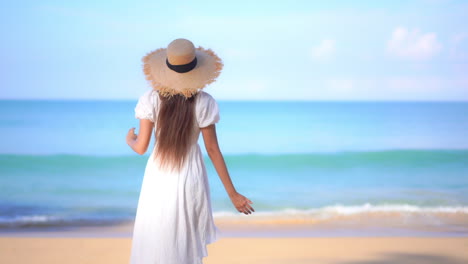 Joven-Viajera-Asiática-Con-Vestido-Blanco-Y-Sombrero-De-Paja-Caminando-Hacia-El-Mar-En-La-Pintoresca-Isla,-Levanta-Los-Brazos-Cuando-Llega-Al-Agua-De-Mar,-Vista-Trasera-En-Cámara-Lenta