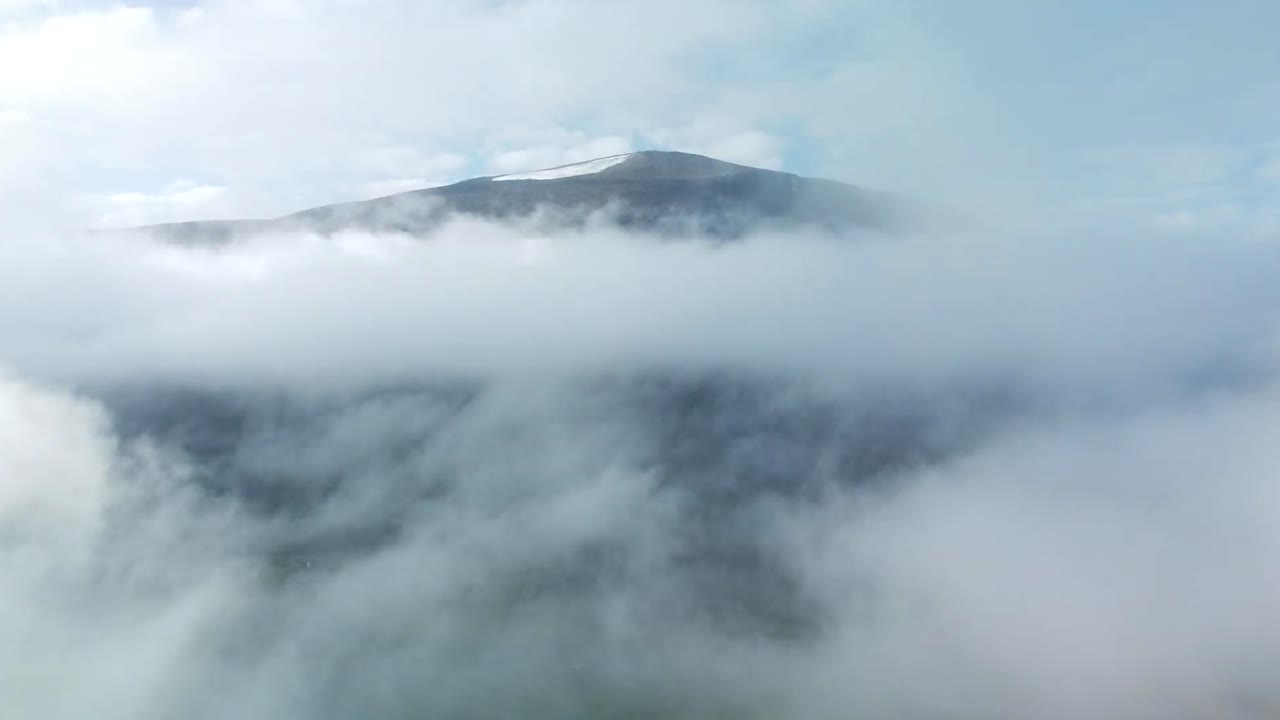 Aerial drone footage flying through white fluffy and thick clouds at a high altitude and approaching Kebnekaise dark black colored mountain in Sweden. Blue sky is visible in the background.